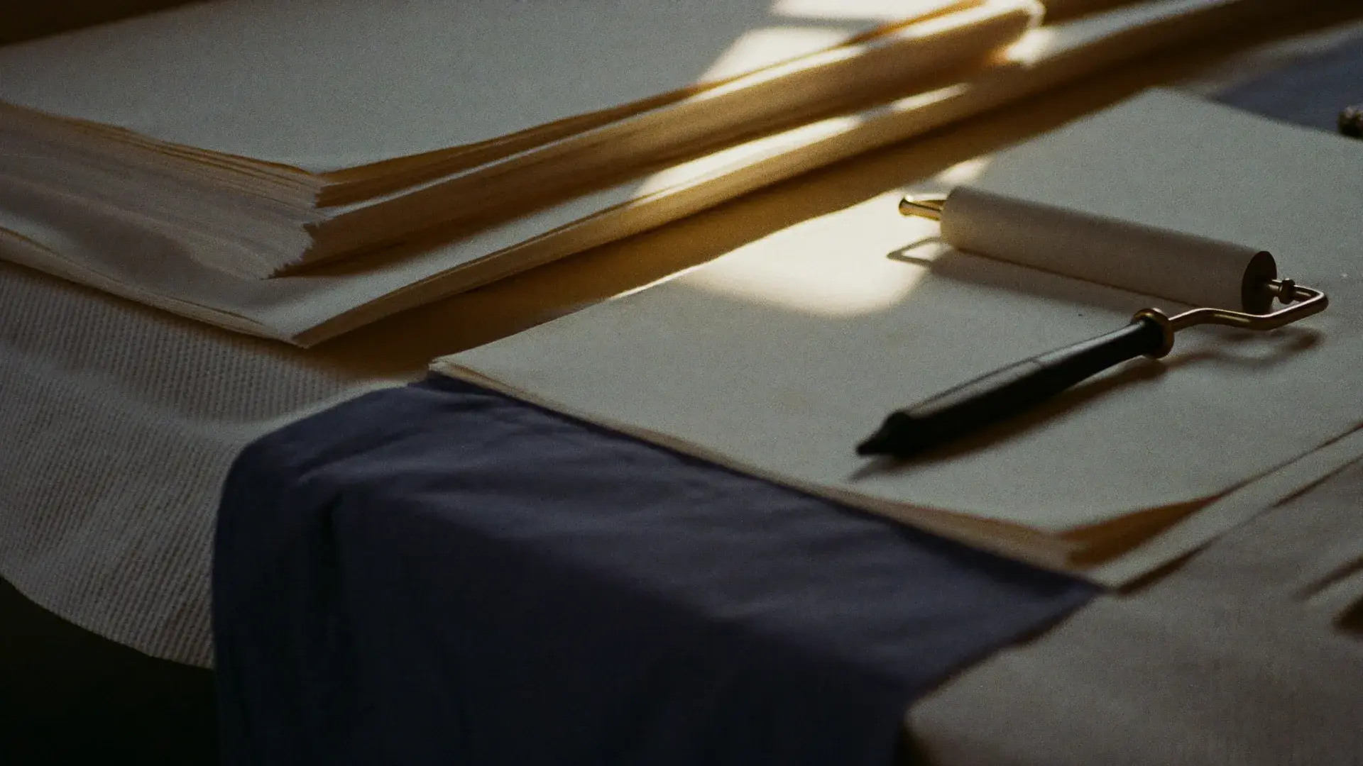 A print studio worktable with stacked paper and an ink roller in warm window light