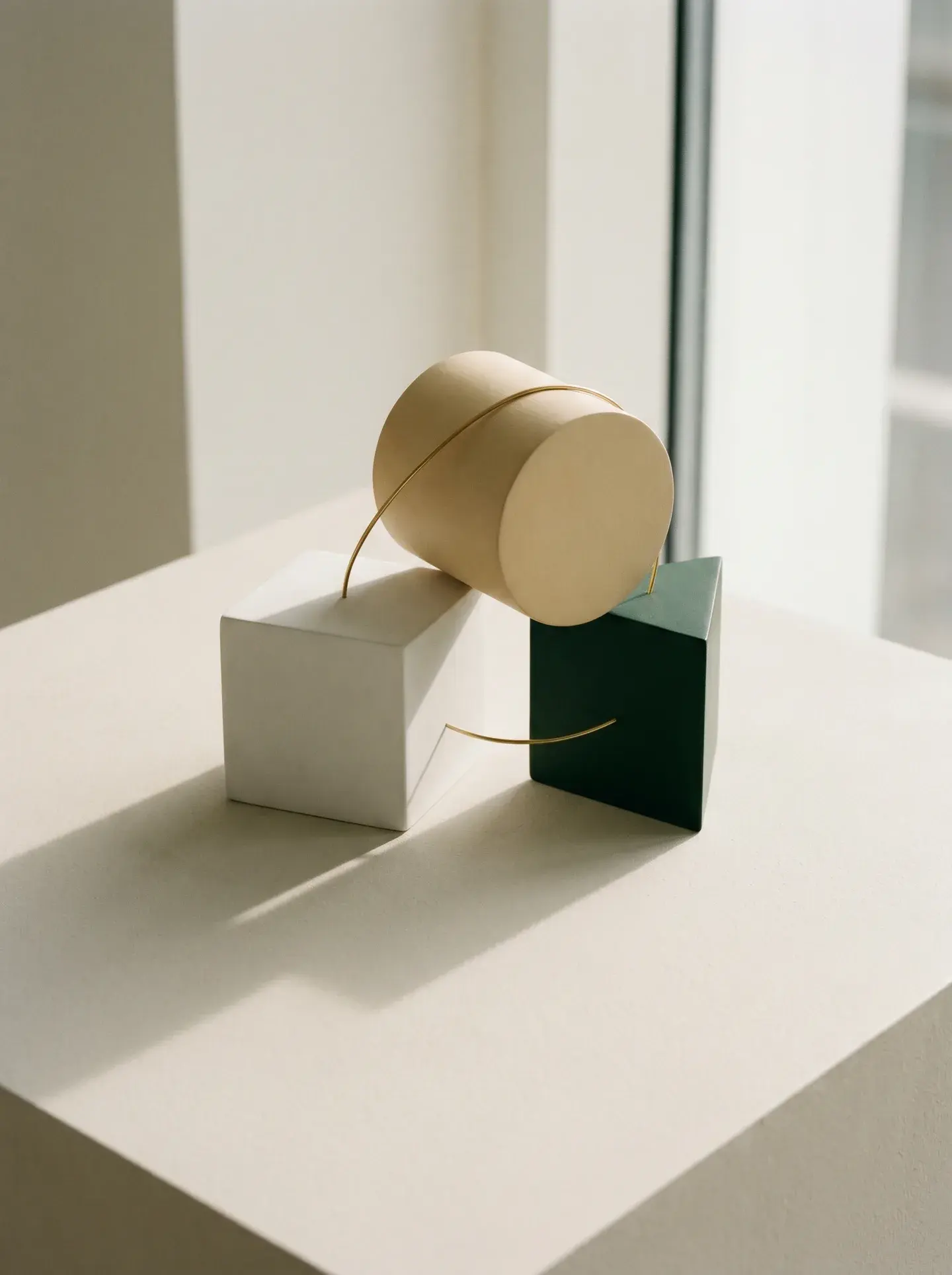 Three interlocking sculptural forms — porcelain cube, parchment cylinder and racing-green prism — joined by a single brass line